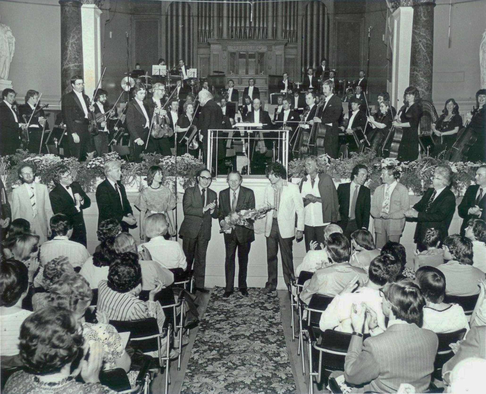 Lennox Berkeley, centre with bouquet, at his 80th birthday concert in Cheltenham Town Hall, 2 July 1983, when the Philharmonia Orchestra conducted by Brian Priestman played Variations on a Theme by Lennox Berkeley. Each of the sixteen one-minute variations on the ‘Reapers’ Chorus’ from the opera Ruth was written by a former pupil. Eleven are shown in this photo (left to right): Brian Chapple, Jonathan Rutherford, Sir John Manduell, Sally Beamish, the late William Mathias, Lennox, Michael Berkeley, Sir John Tavener, Rory Boyle, Richard Stoker, Roy Teed, John McLeod. The others were: the late David Bedford, Christopher Brown, the late Christopher Headington, the late Nicholas Maw.